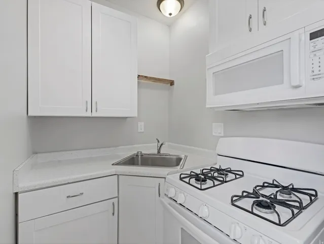 Compact kitchen featuring white cabinetry, a corner stainless steel sink, speckled white quartz countertops, a white gas range, and an over-the-range microwave