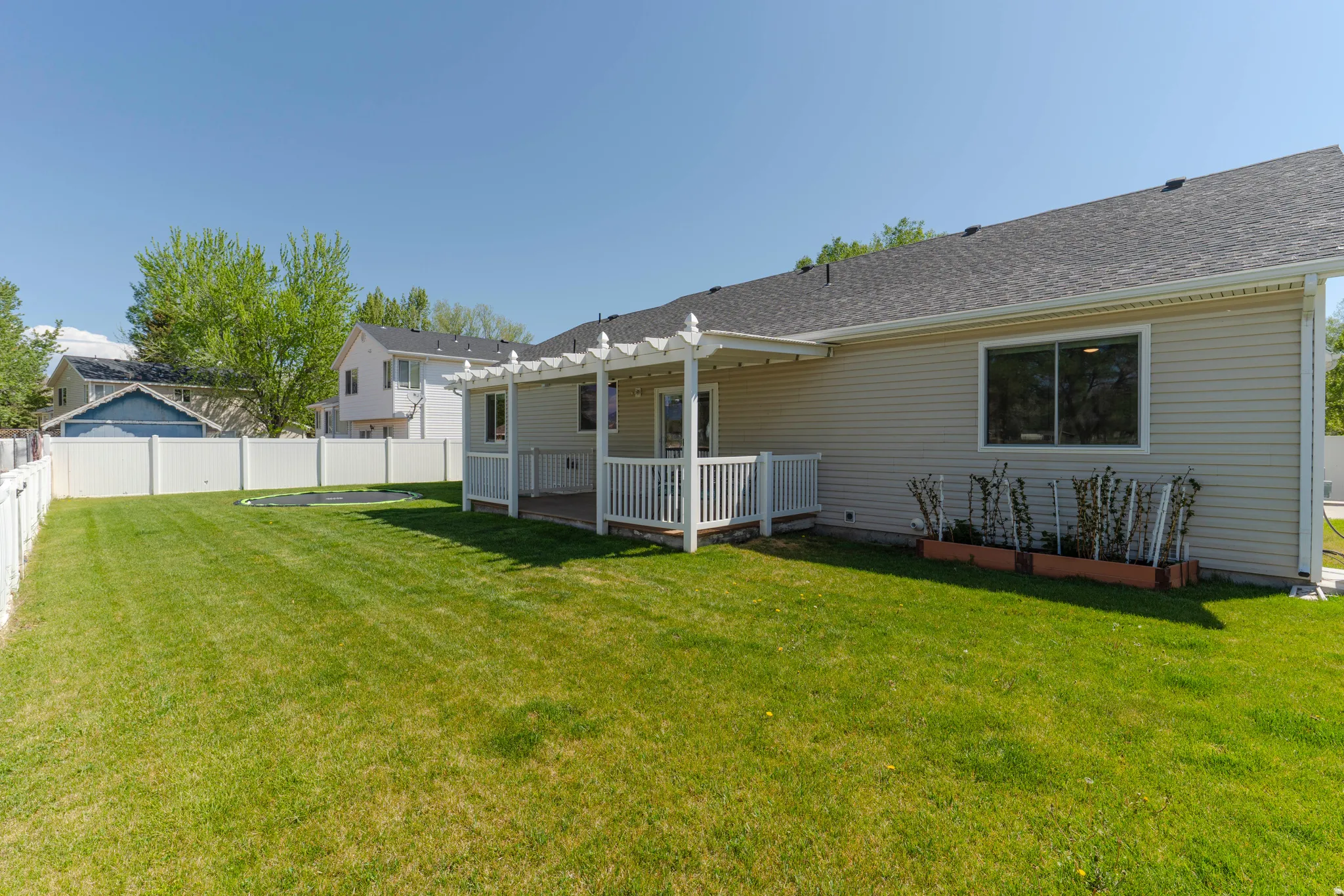 Spacious backyard featuring a manicured lawn, white vinyl fencing, a covered patio with a pergola, and producing raspberry bushes