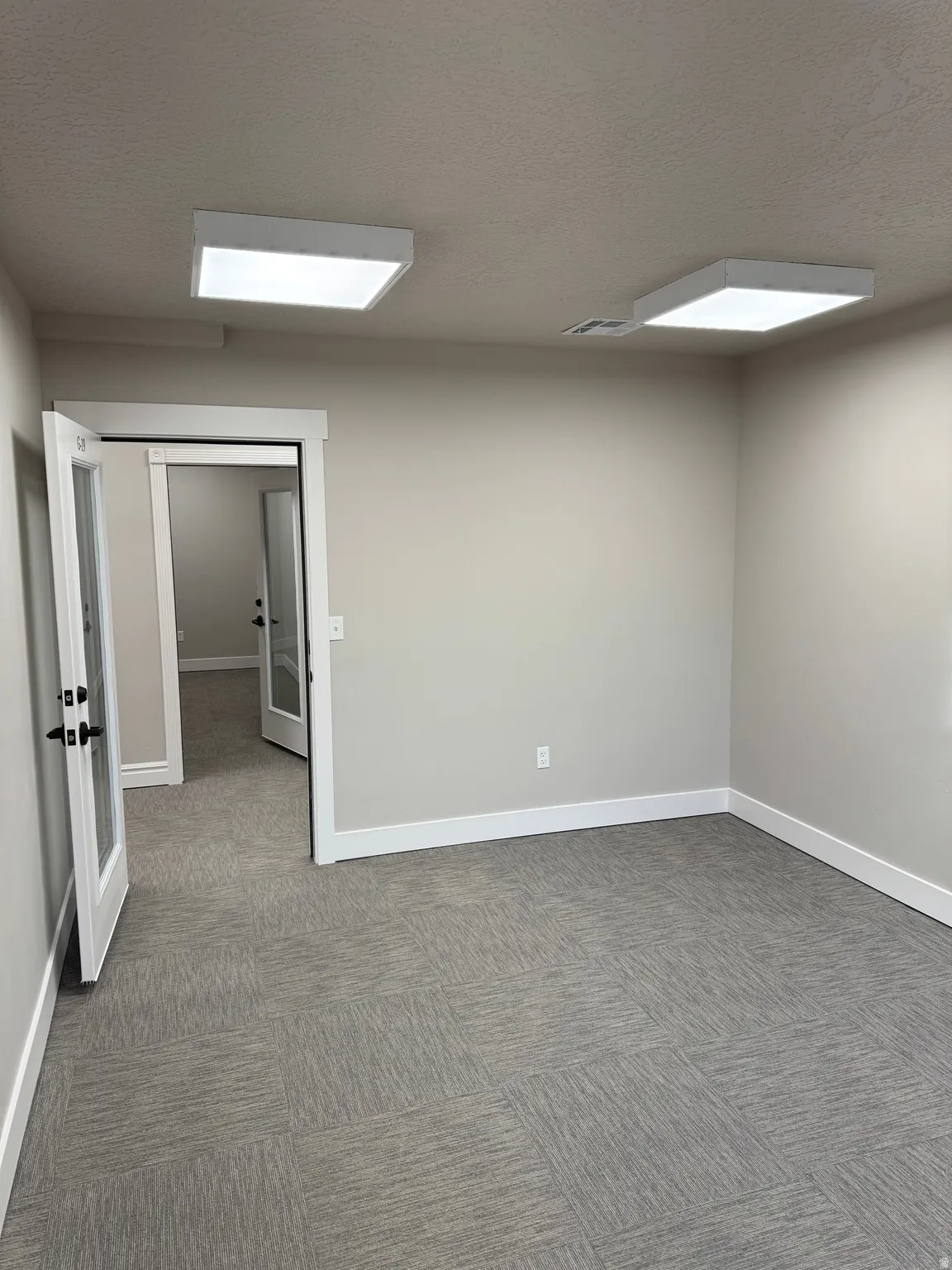 Spacious room featuring gray modular carpet tile, light neutral wall paint, white baseboards, and two square ceiling-mounted light fixtures