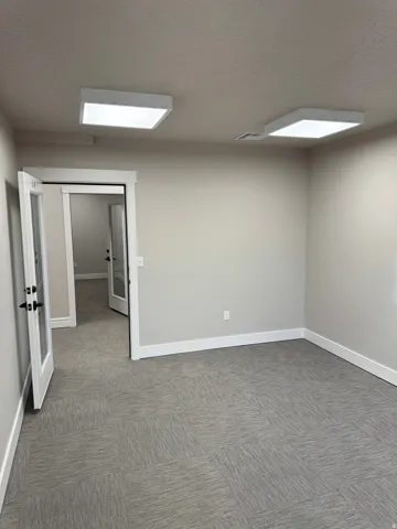 Spacious room featuring gray modular carpet tile, light neutral wall paint, white baseboards, and two square ceiling-mounted light fixtures