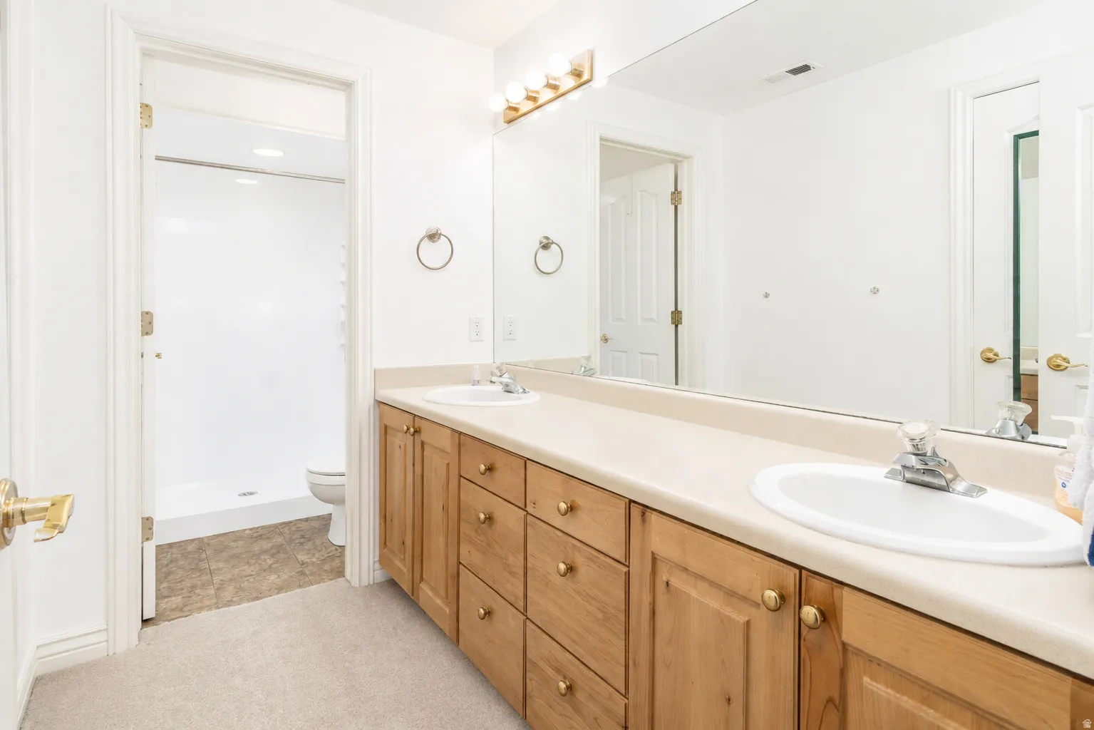 Bathroom featuring a dual vanity with light-tone cabinetry and brass hardware, a light countertop, a large wall-mounted mirror, and overhead vanity lighting