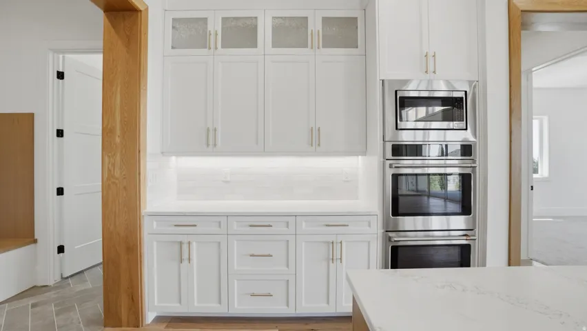 Kitchen featuring white cabinetry with gold-toned hardware, stacked stainless steel appliances, white subway tile backsplash, under-cabinet lighting, and stone countertops