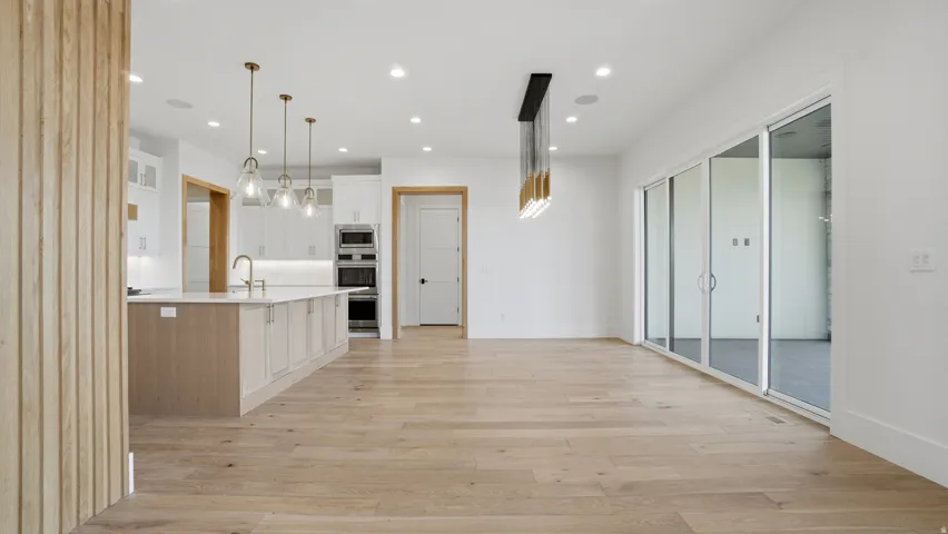 Open-concept interior featuring light wood-finish flooring, a contemporary kitchen with a large island and pendant lighting, white cabinetry, built-in stainless steel appliances, and expansive sliding glass doors