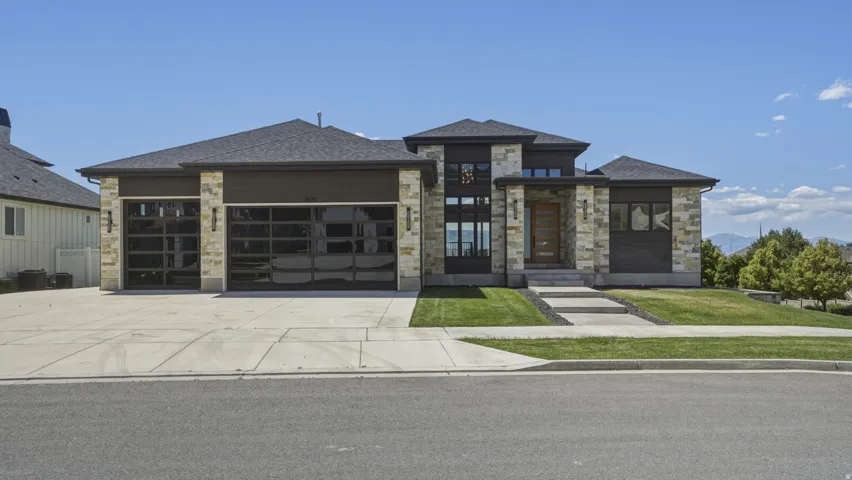 Contemporary residence featuring a stone facade, multi-pane windows, a dark shingle roof, two glass-panel garage doors, and a concrete driveway