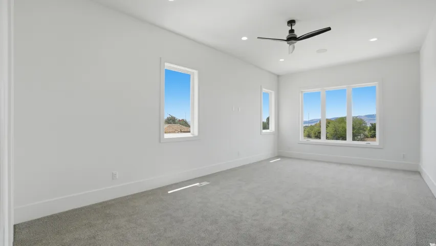 Spacious room featuring light gray carpeting, crisp white walls, recessed ceiling lighting, a modern ceiling fan, and multiple large windows showcasing distant scenery