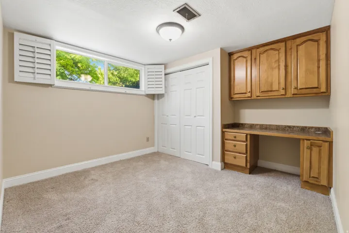 Plush carpeting and neutral wall tones define this room, featuring a built-in wooden desk with upper cabinetry and a contrasting countertop