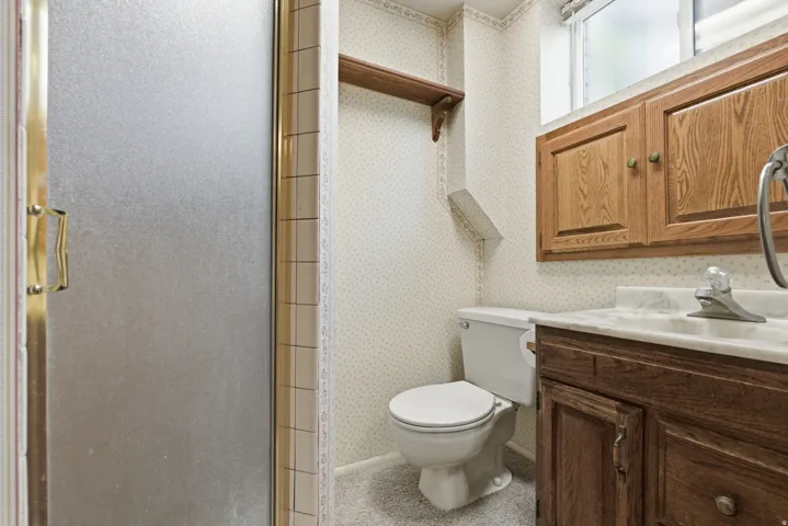 Bathroom featuring a walk-in shower with frosted glass doors, a vanity with a white countertop and wood-finish cabinetry, a toilet, and a window