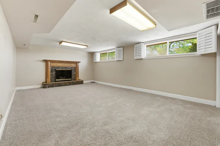 Spacious carpeted room featuring a stone fireplace with a wood mantel, multiple windows with white shutters, and overhead fluorescent lighting