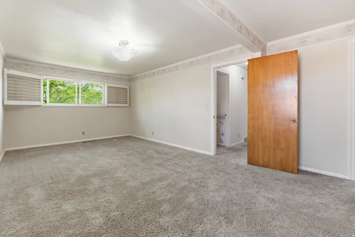 Carpeted room featuring a large window with white plantation shutters, a ceiling-mounted light fixture, and a wood-finish door leading to an additional space