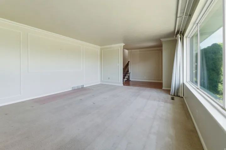 Spacious interior featuring neutral carpeting, extensive paneled wall detailing, crown molding, a large picture window, and wood-finish flooring in the background