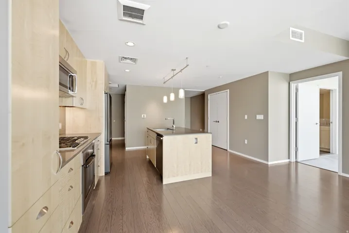 Open-concept kitchen and living area featuring wood-finish flooring, light wood cabinetry, stainless steel appliances, and a central island with a sink and integrated dishwasher