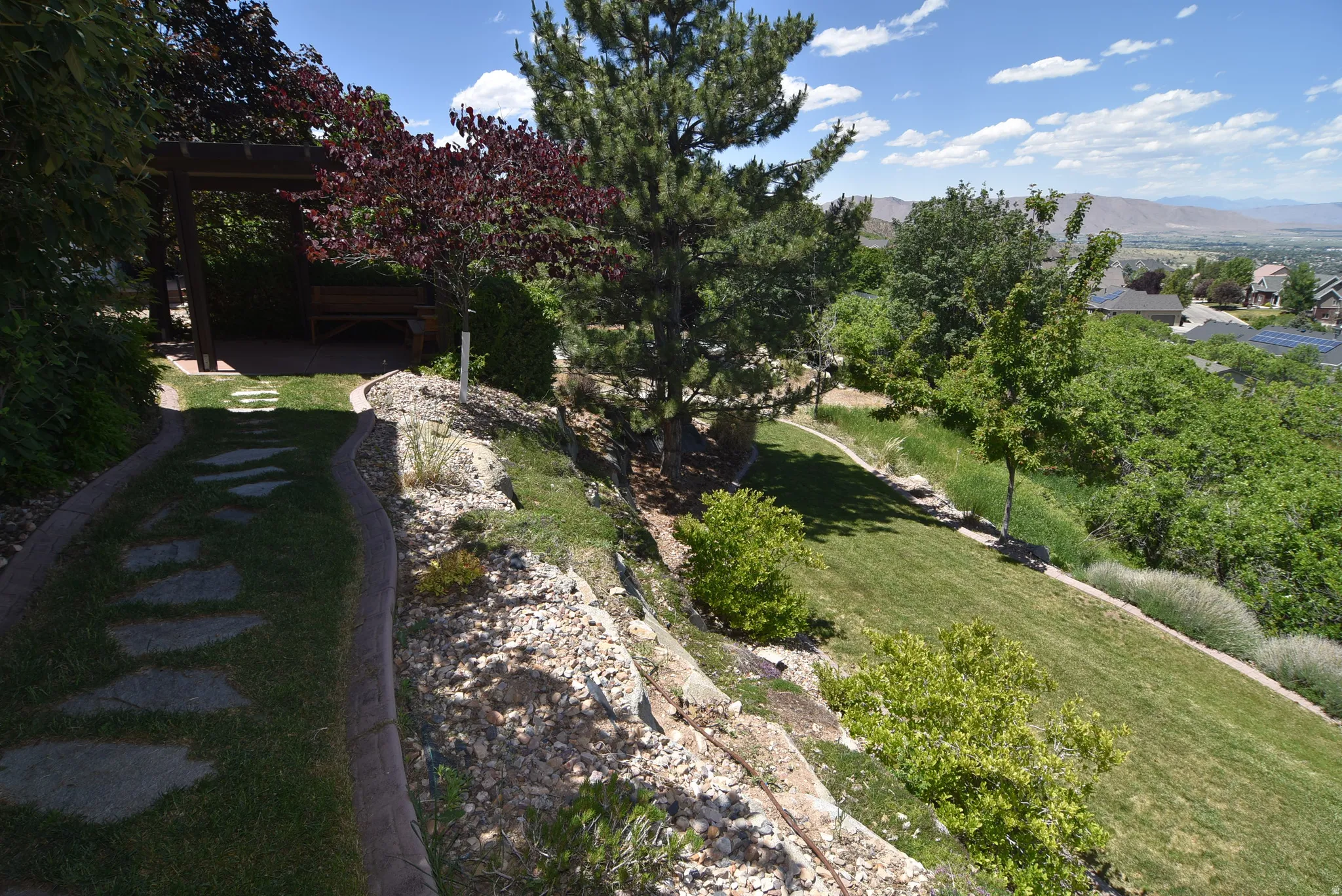 Landscaped terraced backyard featuring a stone pathway, a wooden pergola, mature trees, green lawn, and rock garden elements
