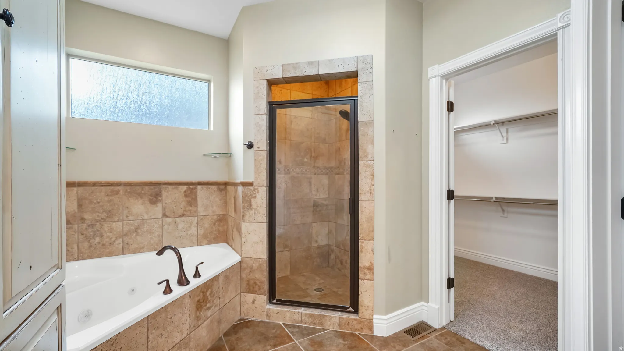 Bathroom featuring a jetted soaking tub with bronze fixtures, a walk-in shower with tile surround and glass enclosure, and an overhead transom window