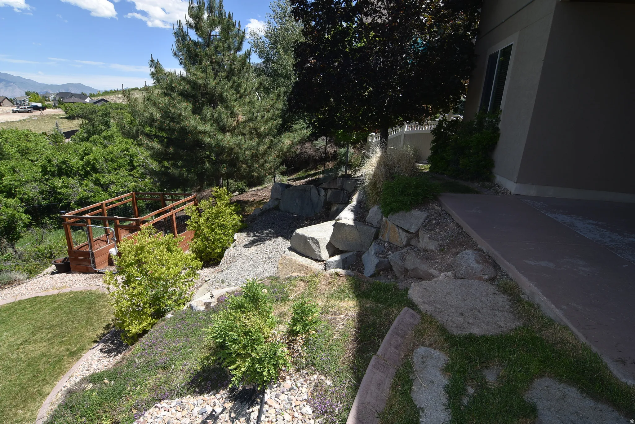 Tiered landscaping featuring a rock retaining wall, mature pine trees, established shrubs, and a built-in fenced structure