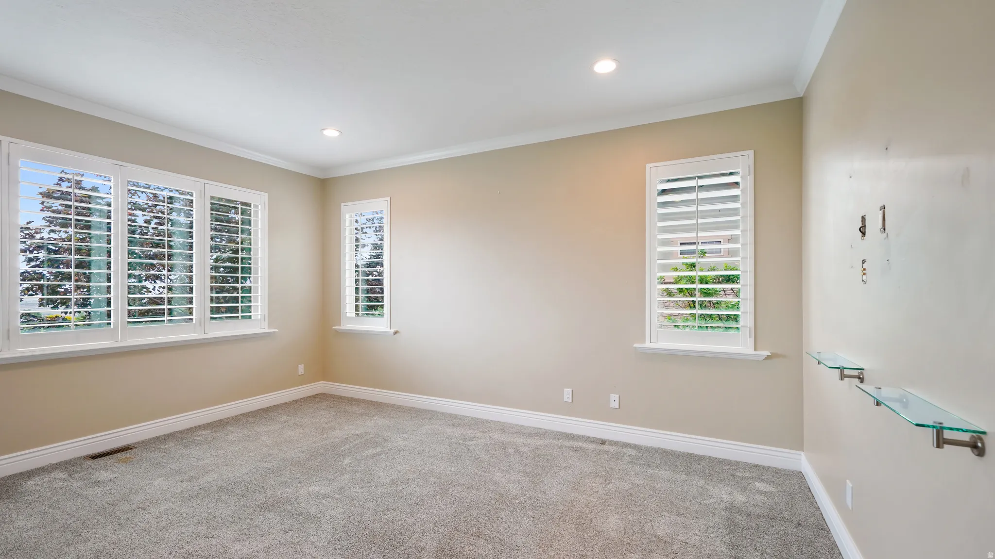 Carpeted interior space featuring multiple windows with plantation shutters, recessed lighting, crown molding, and glass wall shelves