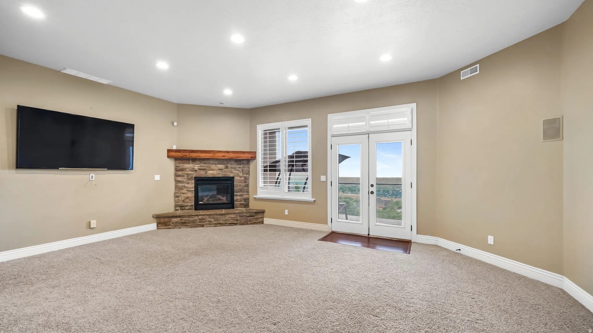 Spacious carpeted room featuring a stone-clad corner fireplace with a wood mantle, recessed lighting, and white French doors opening to an exterior space
