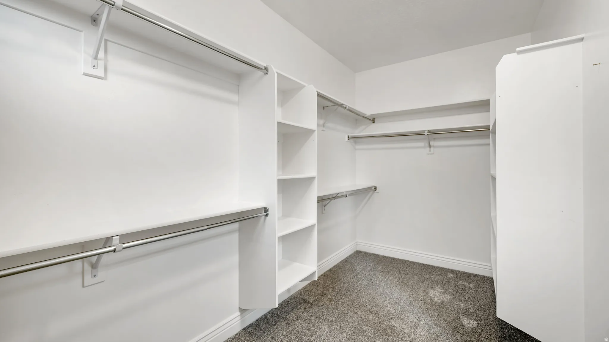 Spacious walk-in closet featuring white built-in shelving, chrome hanging rods, and gray carpet flooring