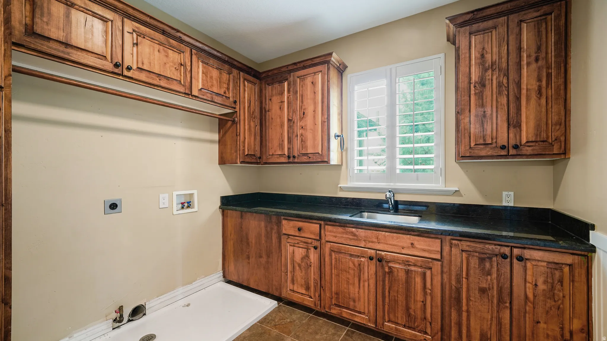 Laundry room featuring natural wood cabinetry, dark countertops, a utility sink with a chrome faucet, and a window with white plantation shutters