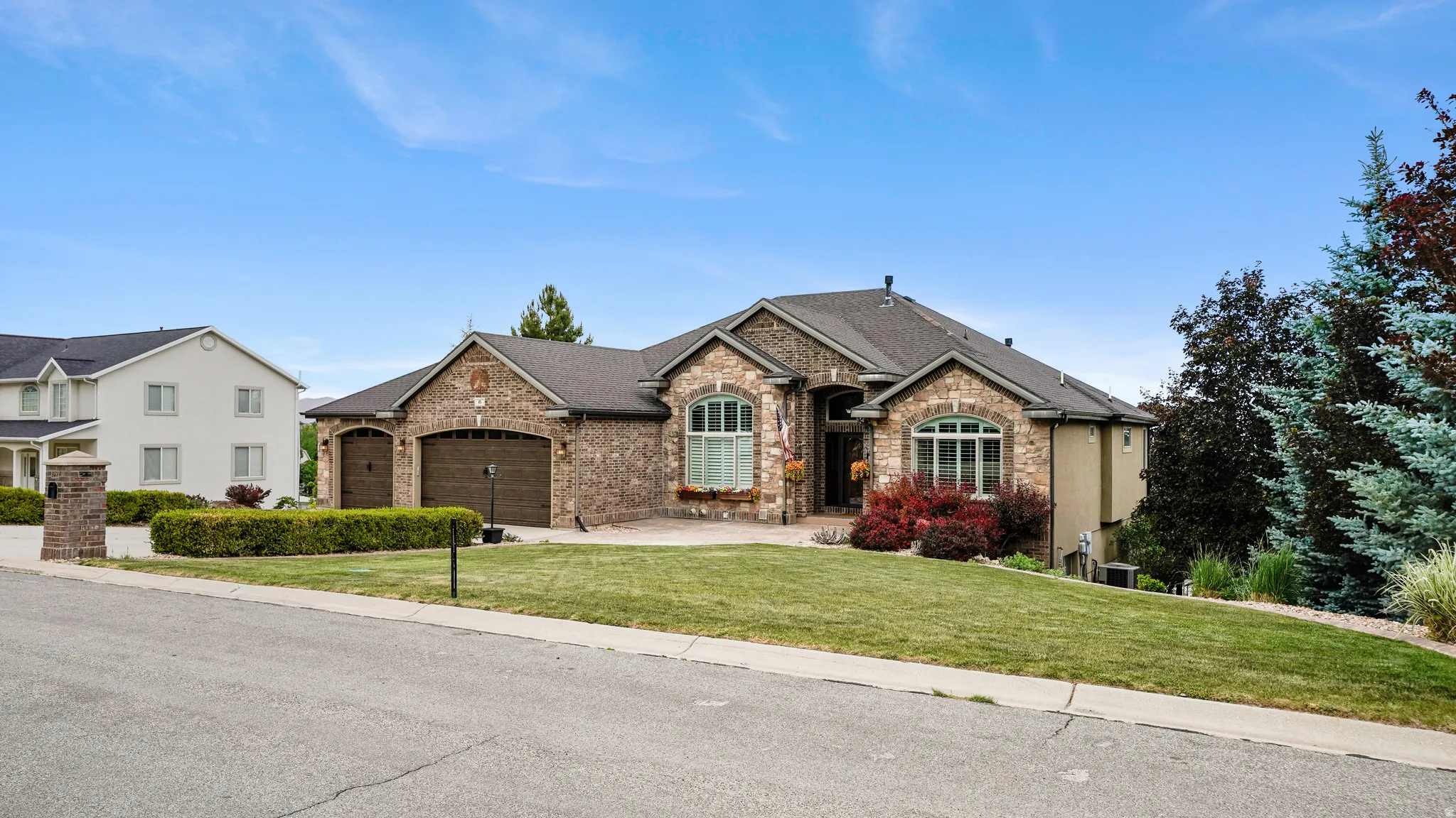 Brick and stucco facade with a multi-car garage, arched windows, and a landscaped front yard featuring mature trees and shrubs