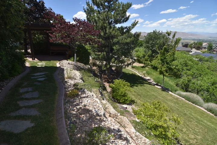 Landscaped terraced backyard featuring a stone pathway, a wooden pergola, mature trees, green lawn, and rock garden elements