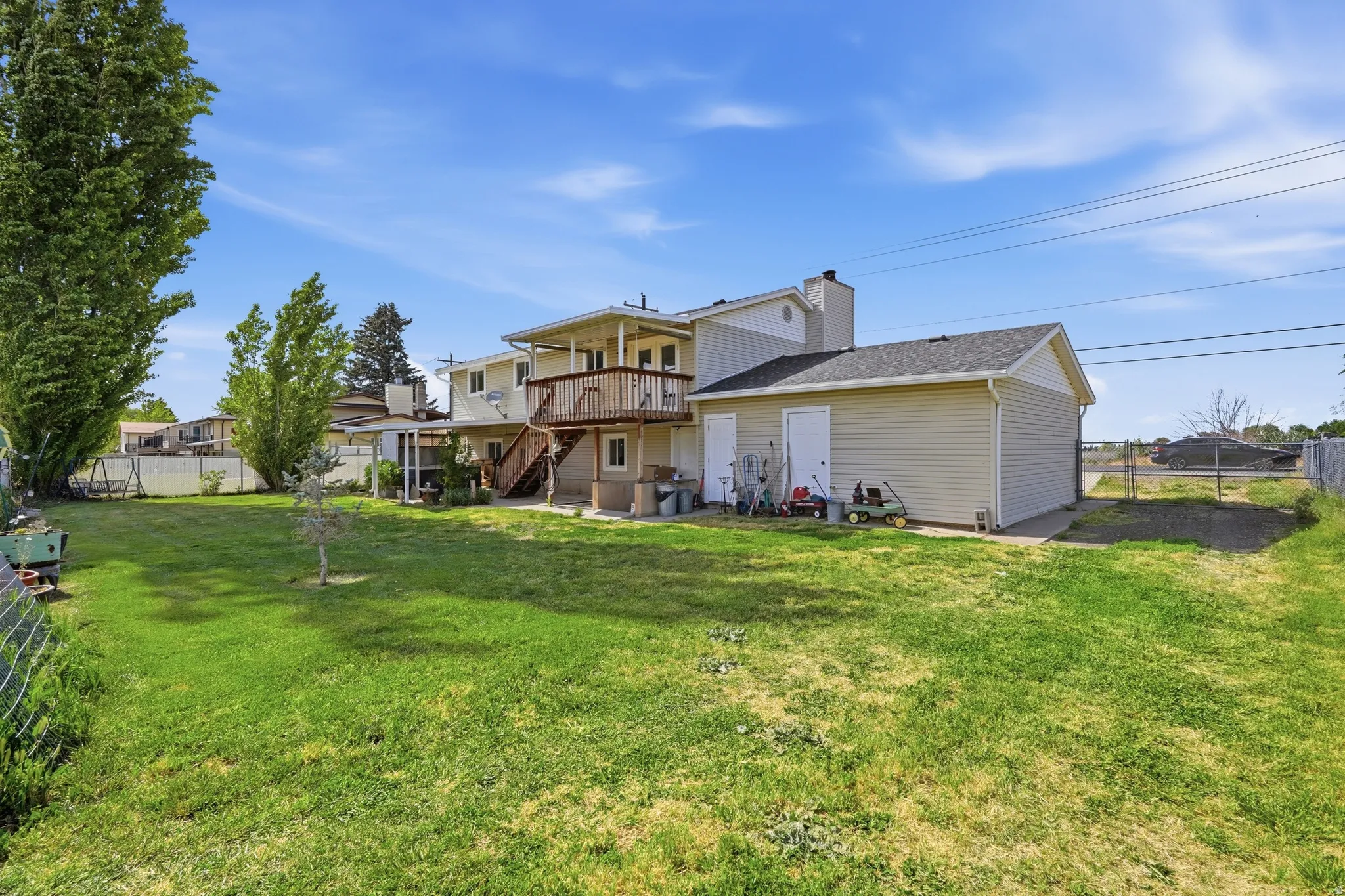 Expansive green lawn with a two-story residence featuring a second-story wooden deck, a ground-level covered patio, and an attached storage shed with two access doors