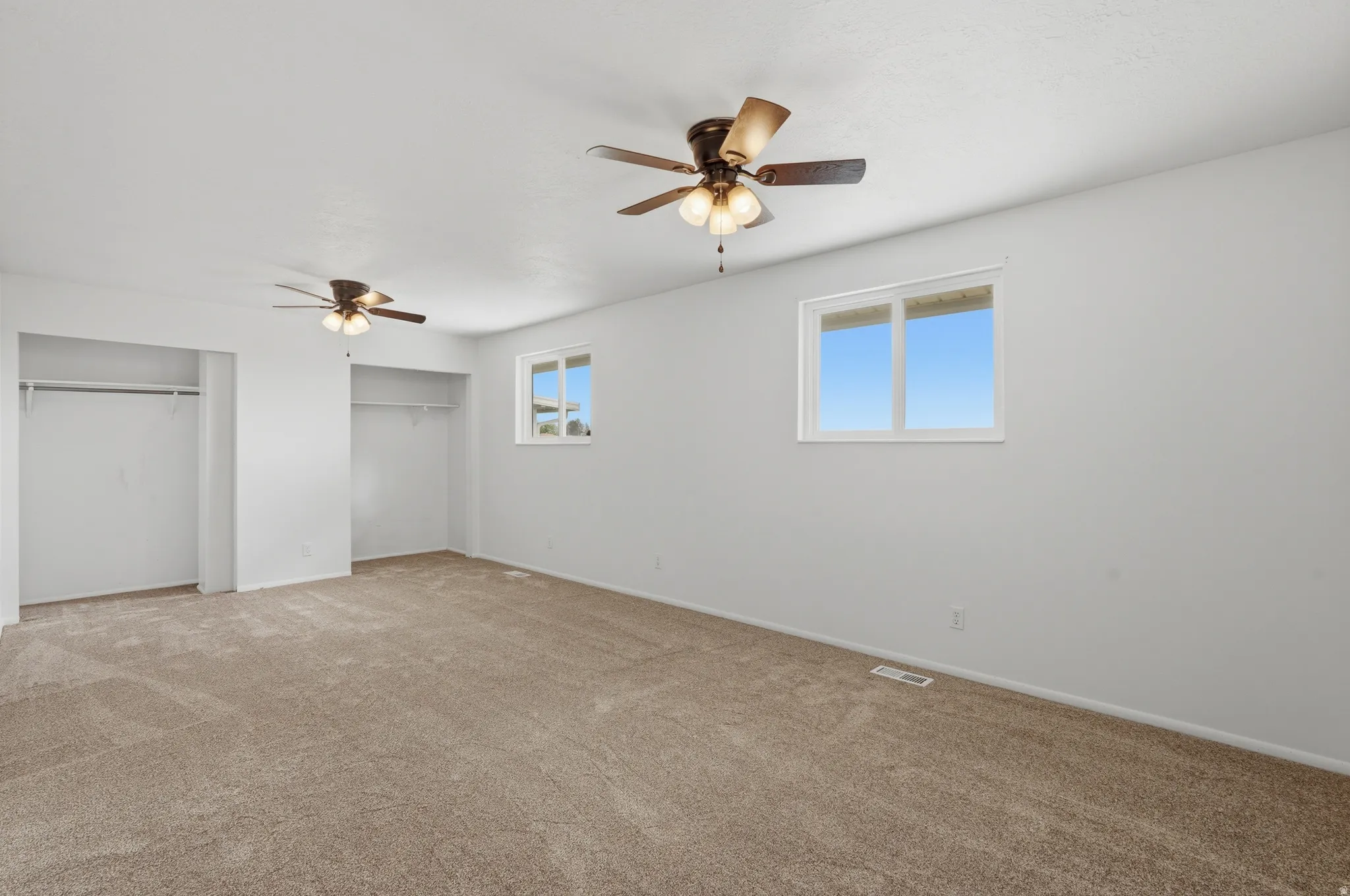 Spacious room featuring neutral carpeting, two ceiling fans with integrated lighting, two windows, and two open closet areas with hanging rods