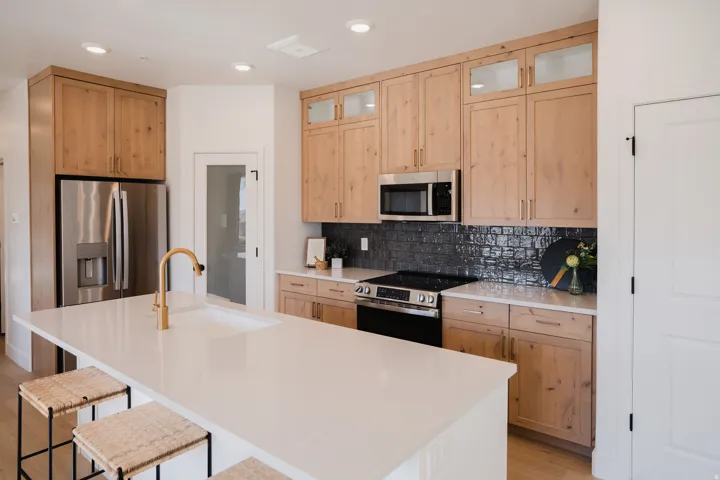 Kitchen featuring a large central island with a built-in sink and gold-finish faucet, light wood cabinetry, stainless steel appliances, and a dark tile backsplash