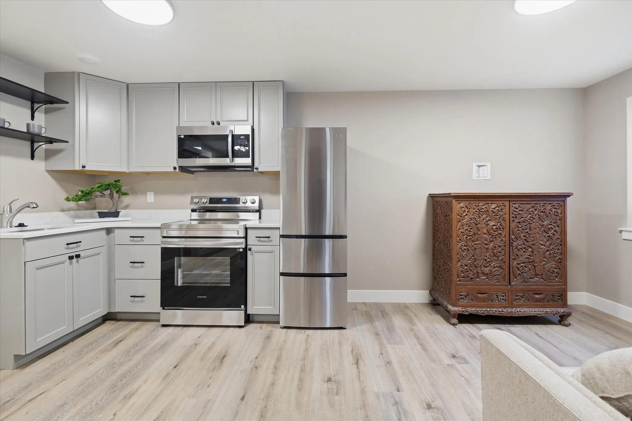 Apartment -Modern kitchen area featuring light gray cabinetry, stainless steel appliances, open shelving, and wood-finish flooring