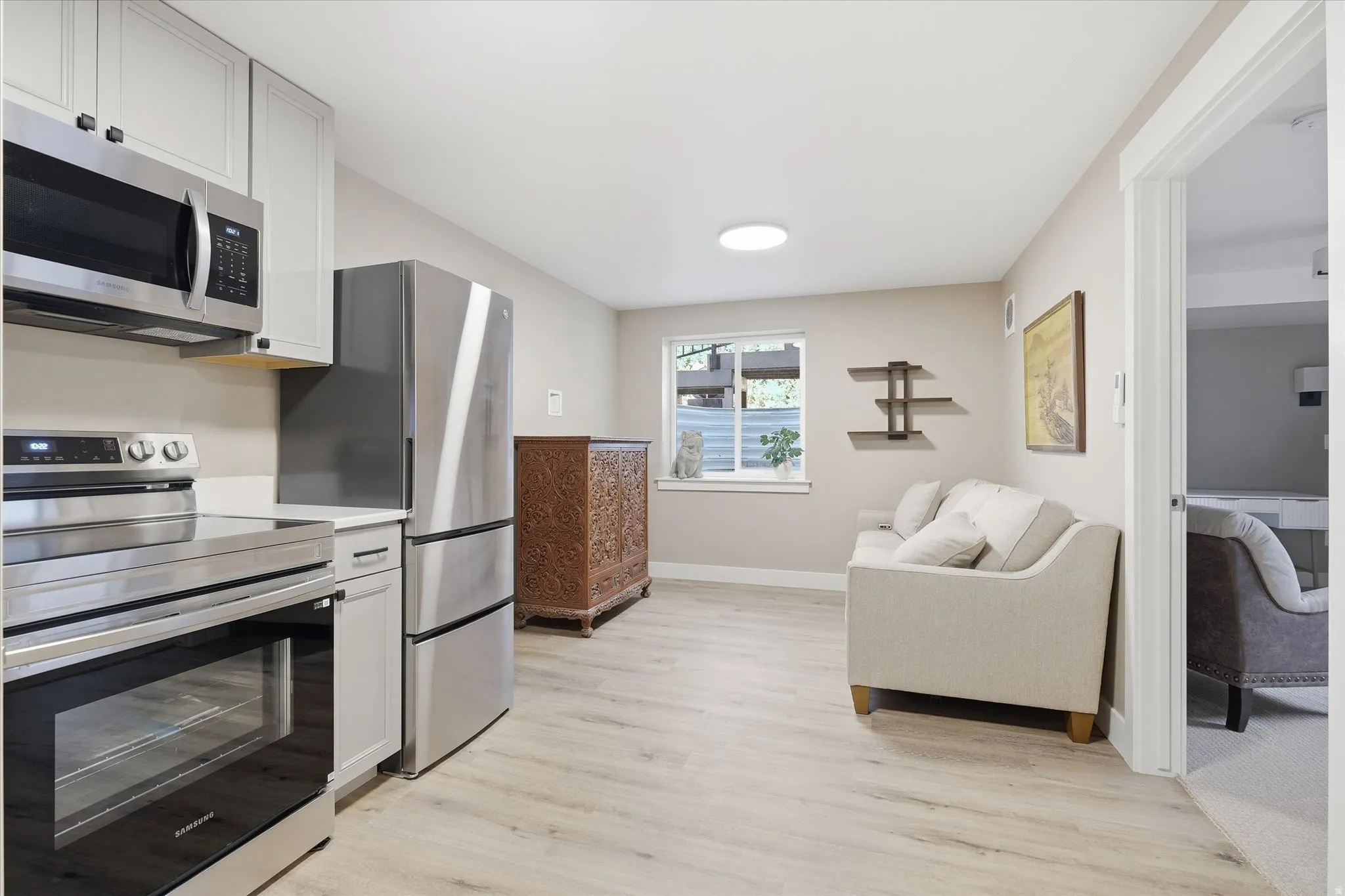 Apartment -Kitchenette and living area featuring light wood-finish flooring, white cabinetry, and stainless steel appliances including a refrigerator, range, and microwave
