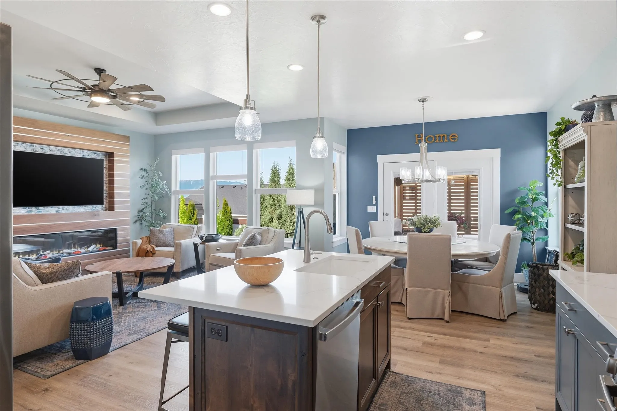 Open-concept living area featuring a kitchen island with seating, wood-finish flooring, a contemporary linear fireplace with stacked stone surround, multiple windows, and recessed lighting