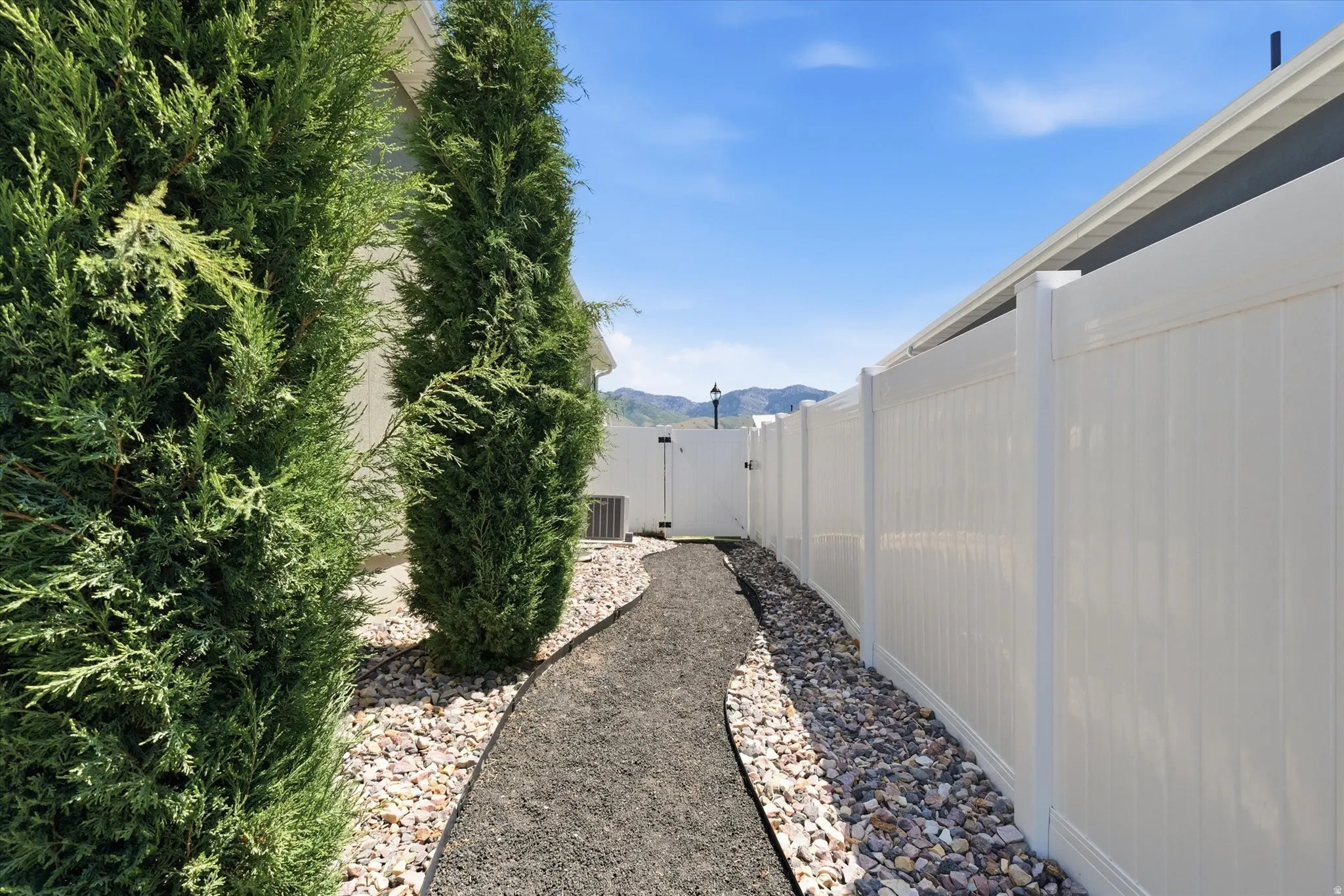 Landscaped side yard featuring a white privacy fence, an air conditioning unit, and a gravel pathway