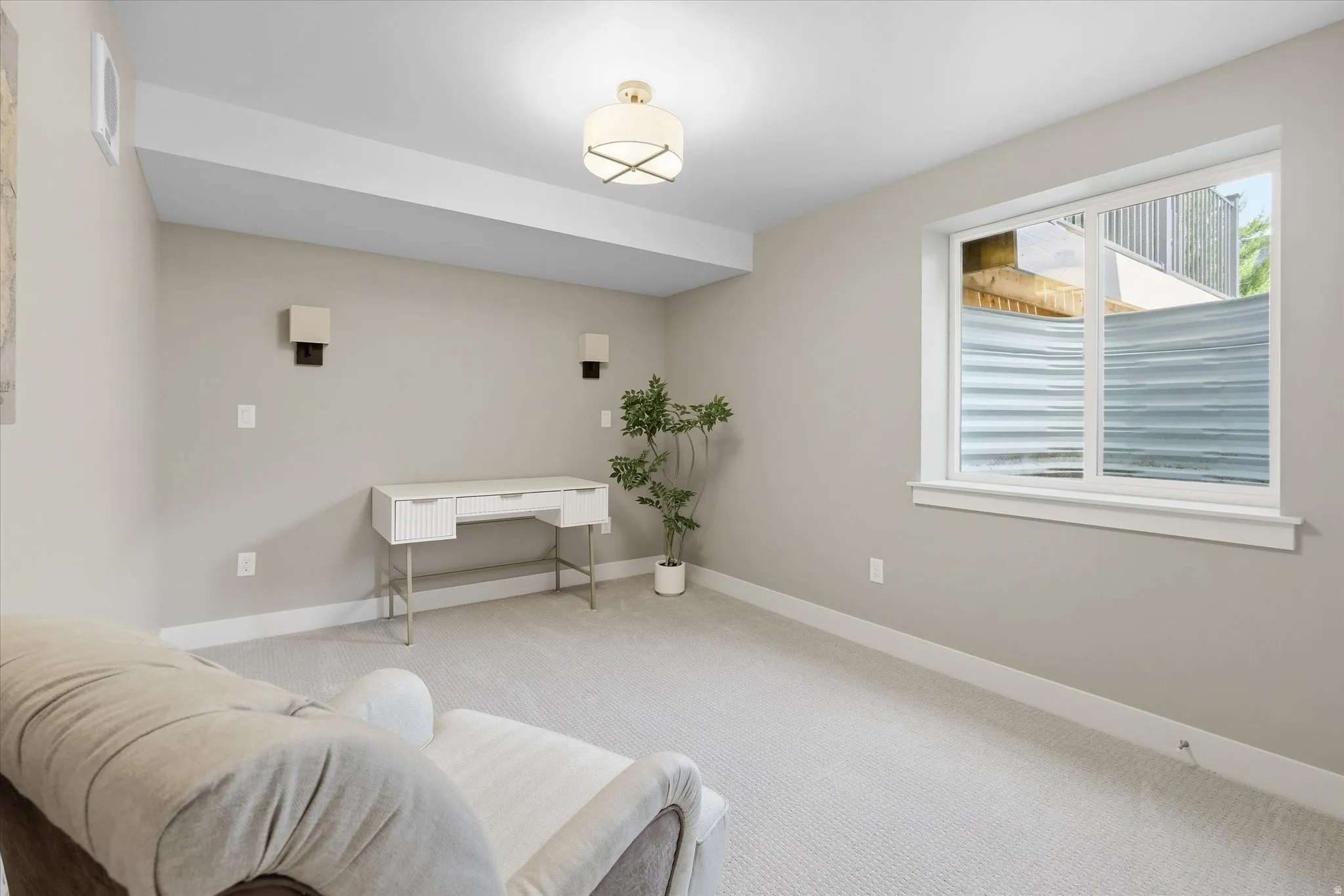 Apartment - Carpeted room featuring a white-framed window, two wall-mounted sconces, and a contemporary flush-mount ceiling light