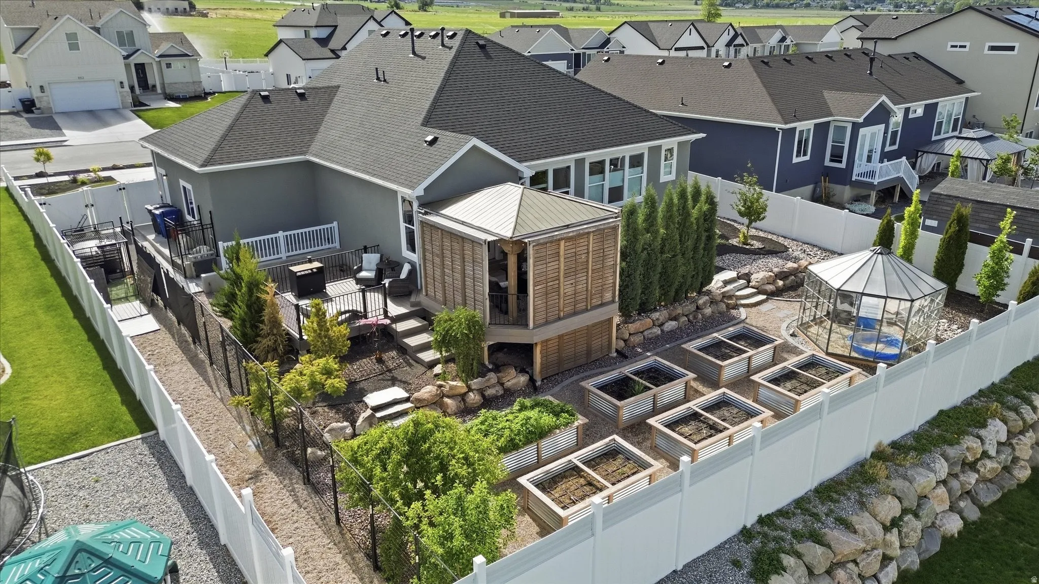 Fully fenced rear yard featuring a multi-level deck, a pergola structure, a glass greenhouse, multiple raised garden beds, and an extensive rock retaining wall