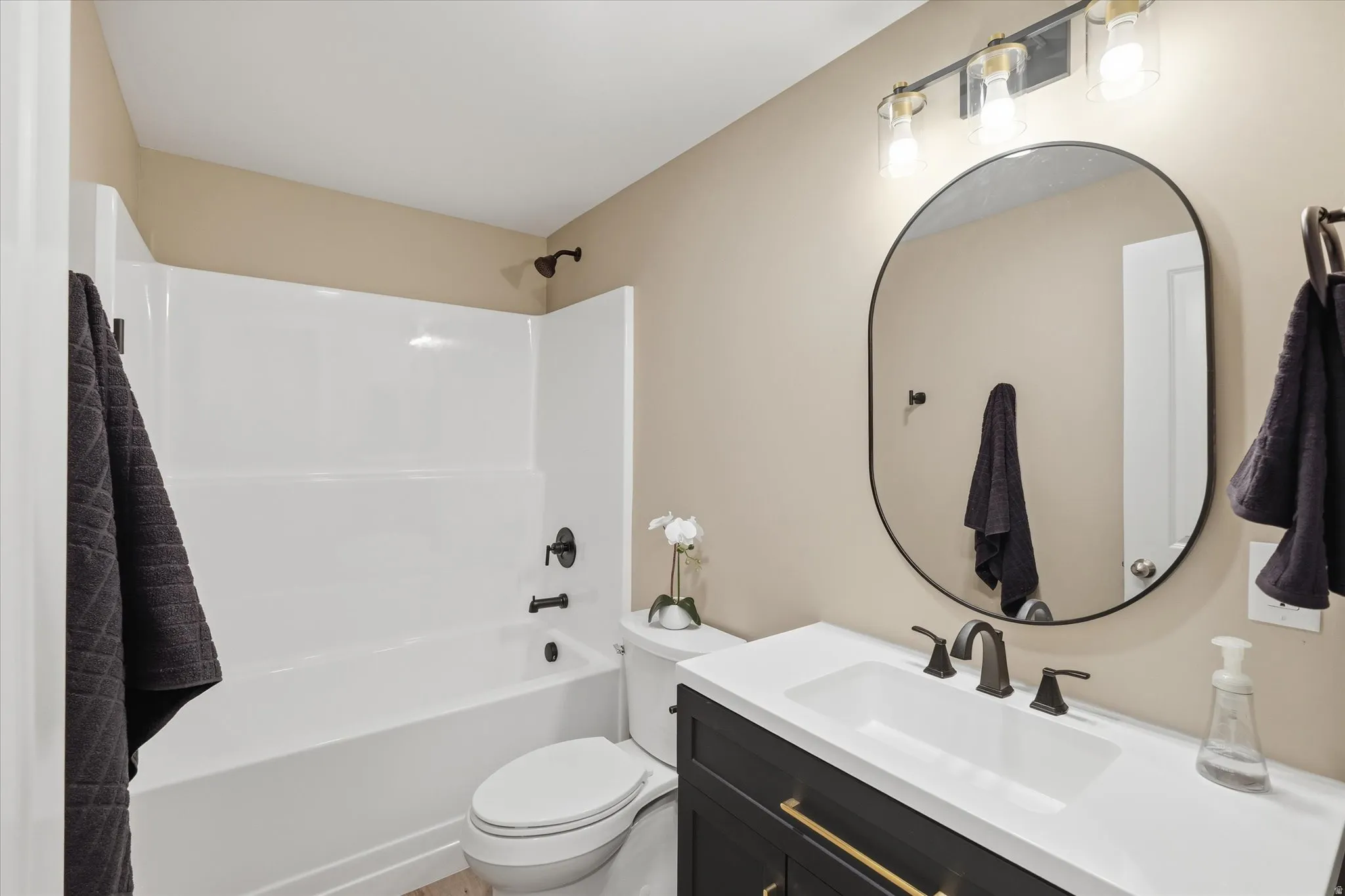 Bathroom featuring a white bathtub and shower surround, a dark vanity with gold-tone hardware, a white countertop with an integrated sink, a black oval mirror, and a three-light fixture with clear glass shades