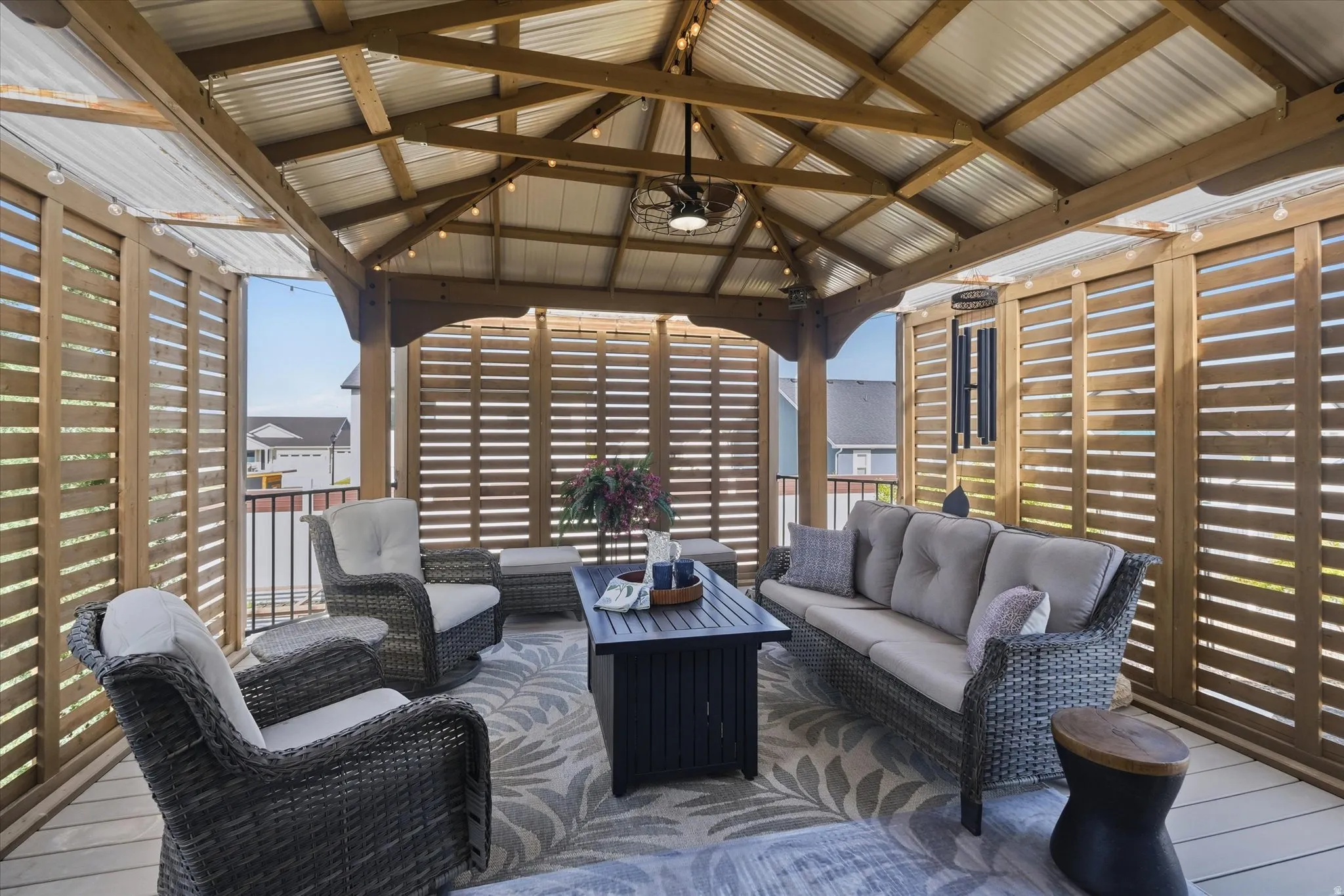 Outdoor gazebo featuring a corrugated metal roof, exposed wood beams, slatted wood privacy walls, and strung overhead lighting