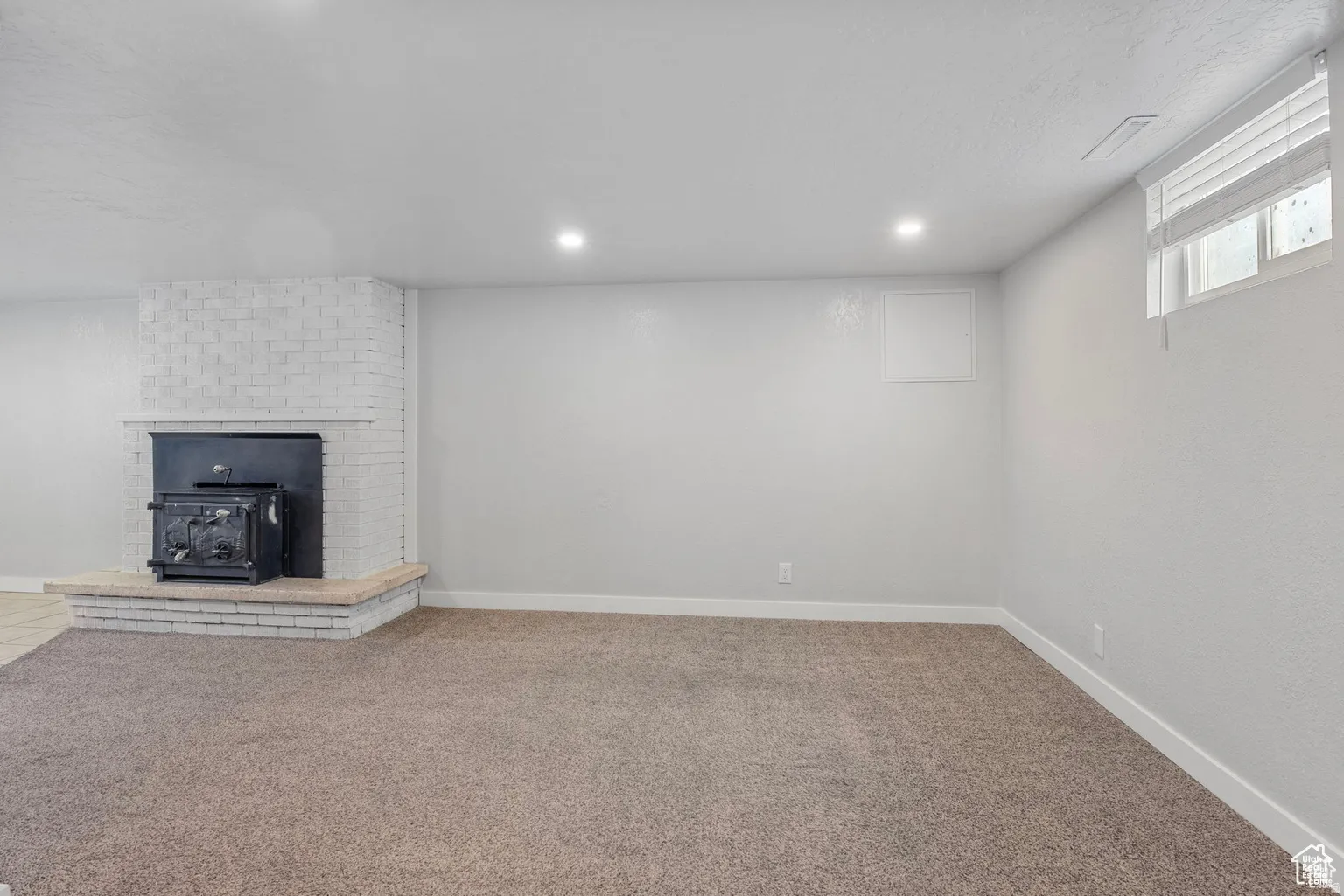 Carpeted interior space featuring a white brick fireplace with a black wood-burning stove insert, recessed lighting, and a white framed window with horizontal blinds