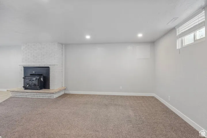Carpeted interior space featuring a white brick fireplace with a black wood-burning stove insert, recessed lighting, and a white framed window with horizontal blinds