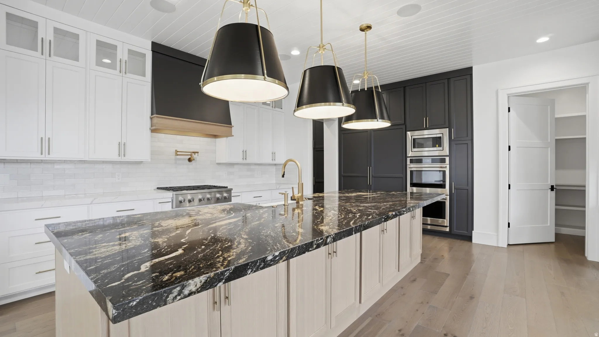 Kitchen featuring a large island with a patterned countertop, brass-toned fixtures, white shaker-style cabinetry, dark cabinetry surrounding built-in appliances, and light wood-finish flooring