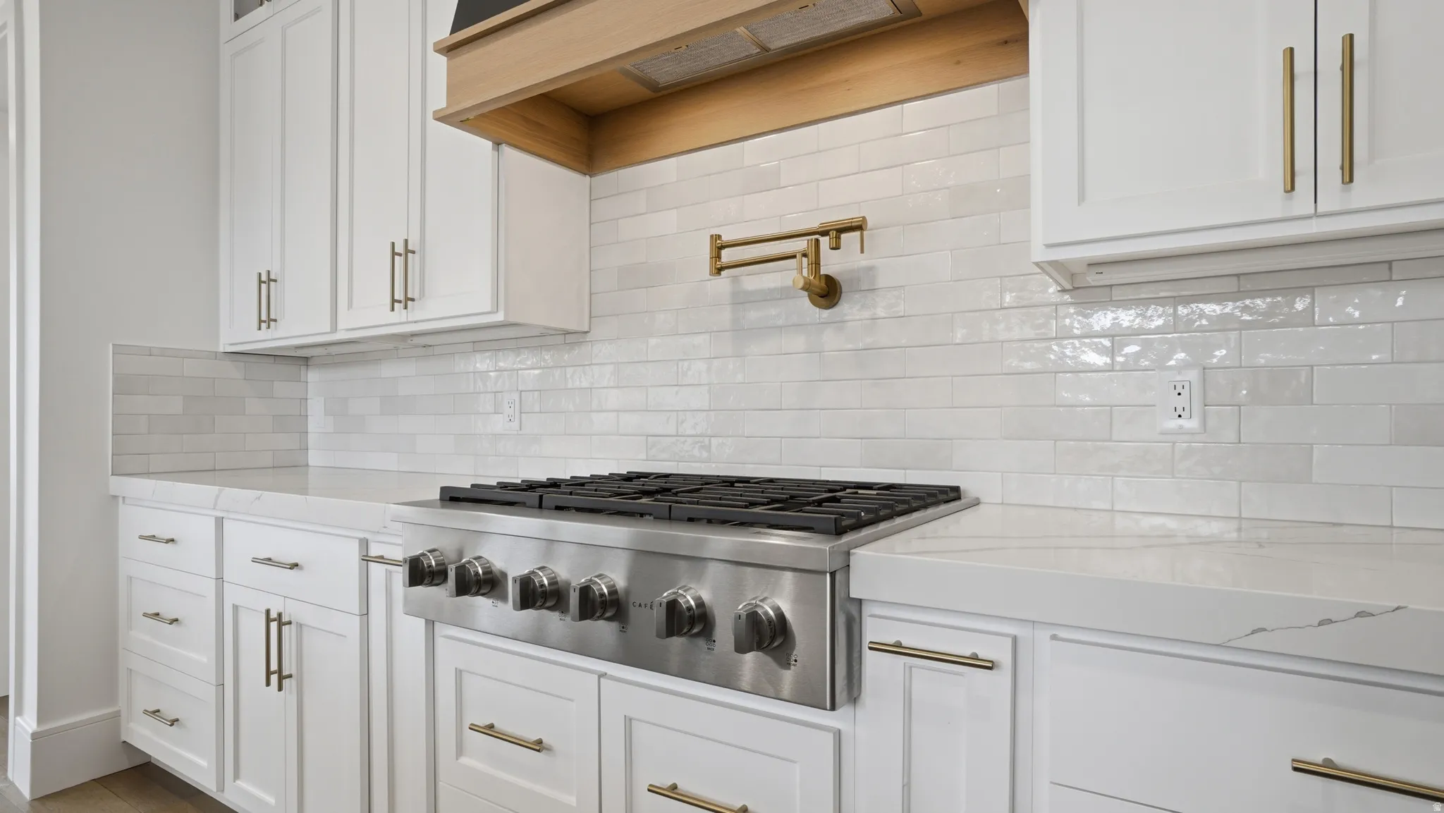 Gourmet kitchen featuring a stainless steel range, a brass pot filler, white shaker-style cabinetry with brass hardware, a light wood-finish range hood, and a white subway tile backsplash