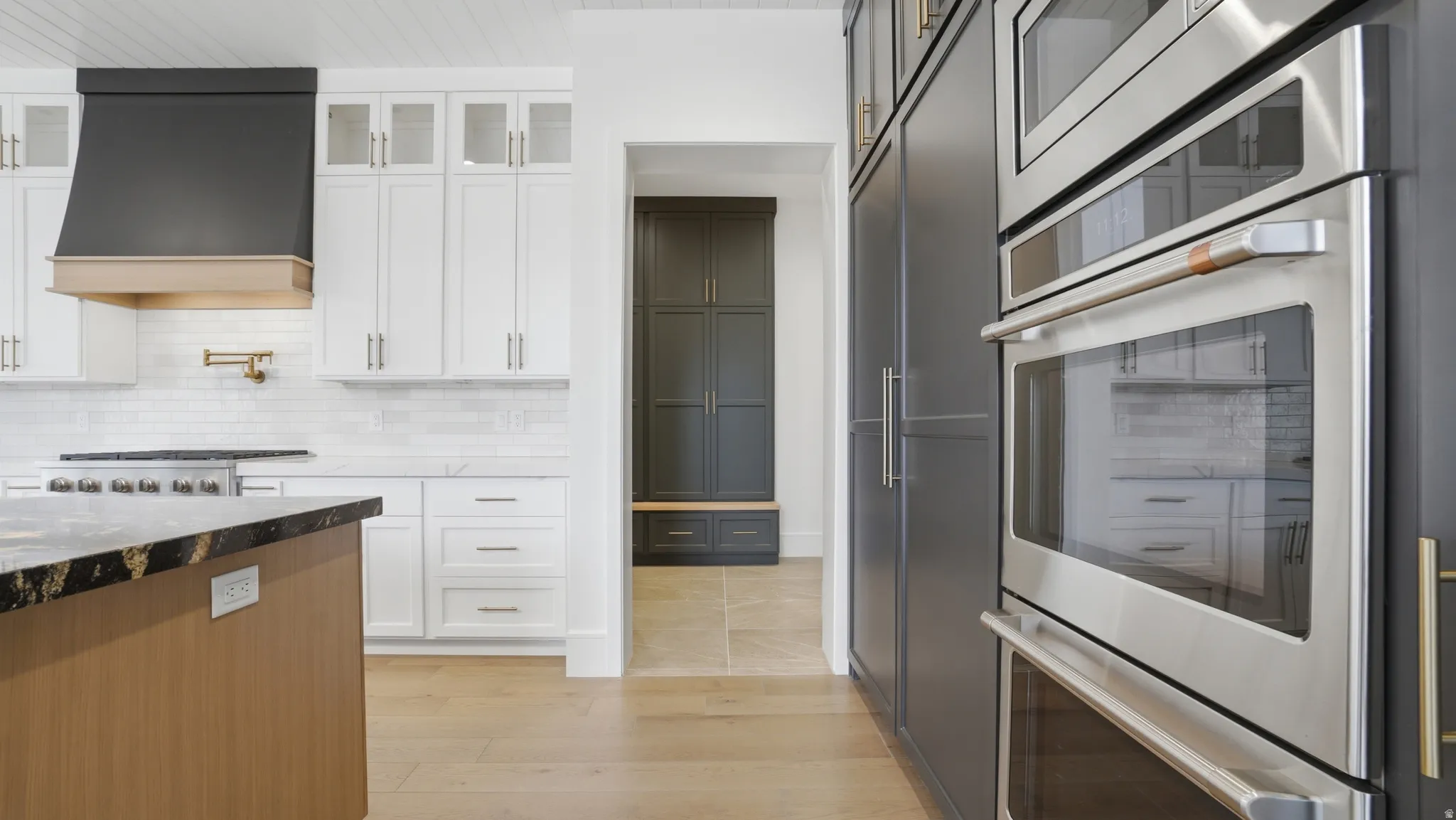 Gourmet kitchen featuring a built-in double oven, full-height dark cabinetry, a light wood-finish island with a dark countertop, white upper and lower cabinetry, and a black range hood with a wood-finish accent