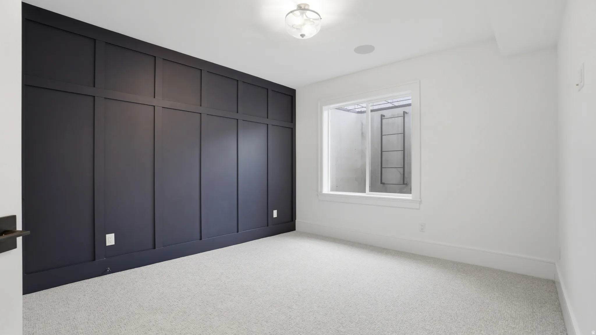 Accent wall with dark wainscoting and trim, light gray textured carpet, recessed ceiling speaker, ceiling-mounted light fixture, and white window trim