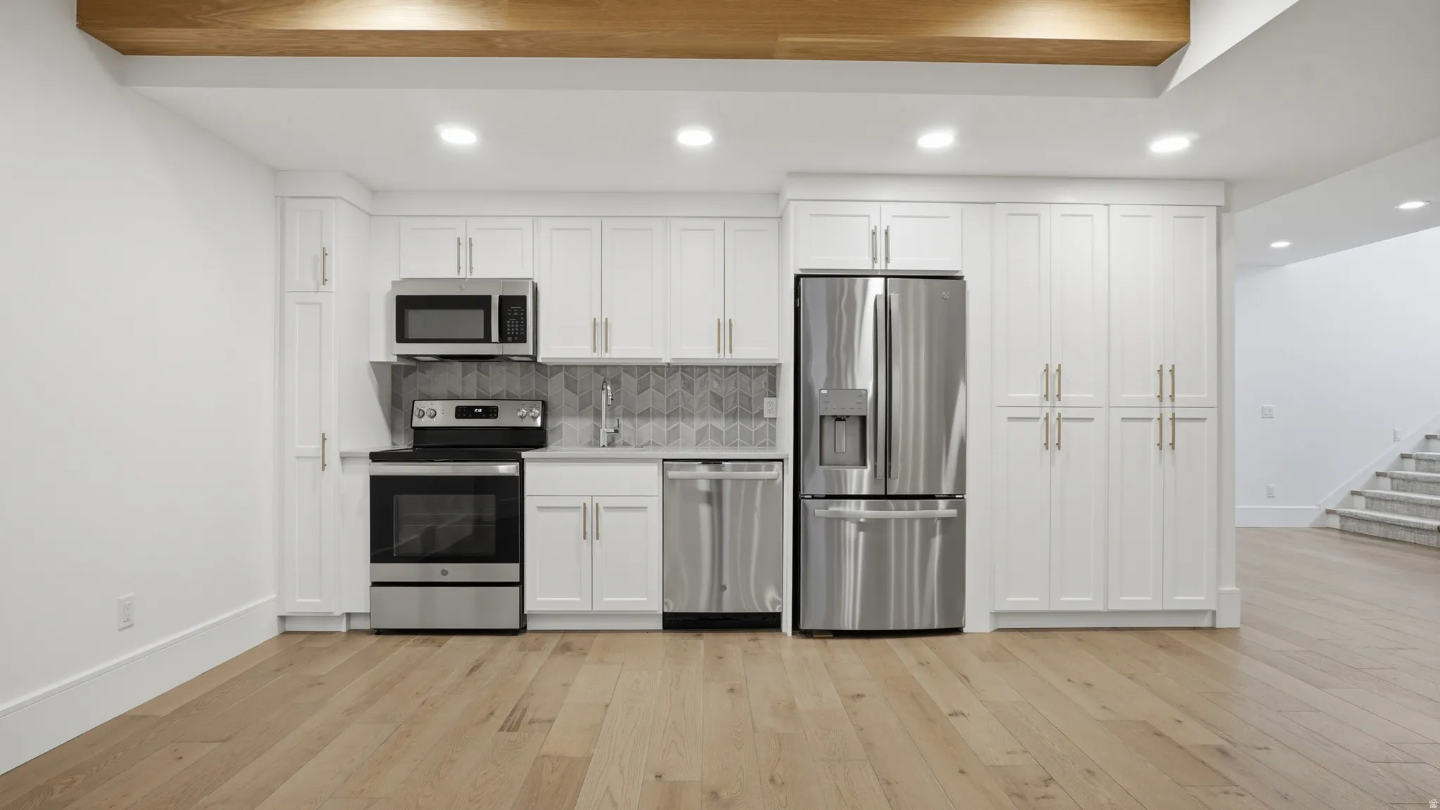 Kitchenette featuring stainless steel appliances, white cabinetry with gold-tone hardware, a gray herringbone tile backsplash, and light wood-finish flooring