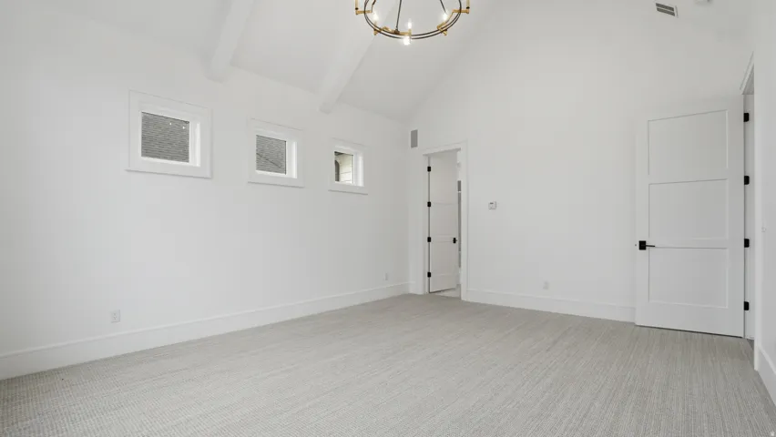 Spacious room featuring vaulted ceilings with exposed beams, light-colored carpeting, multiple windows, and a modern chandelier