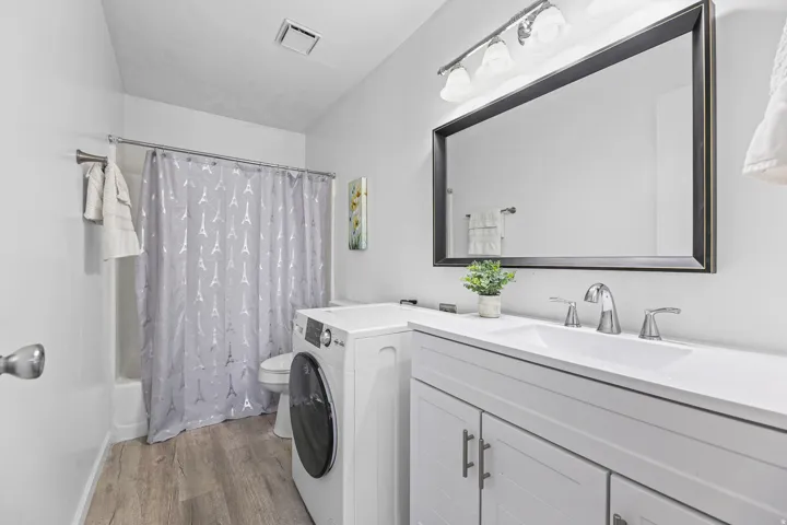 Contemporary bathroom featuring a single vanity with storage, a rectangular sink with chrome fixtures, a large framed mirror, vinyl wood-finish flooring and an all in one washer/dryer