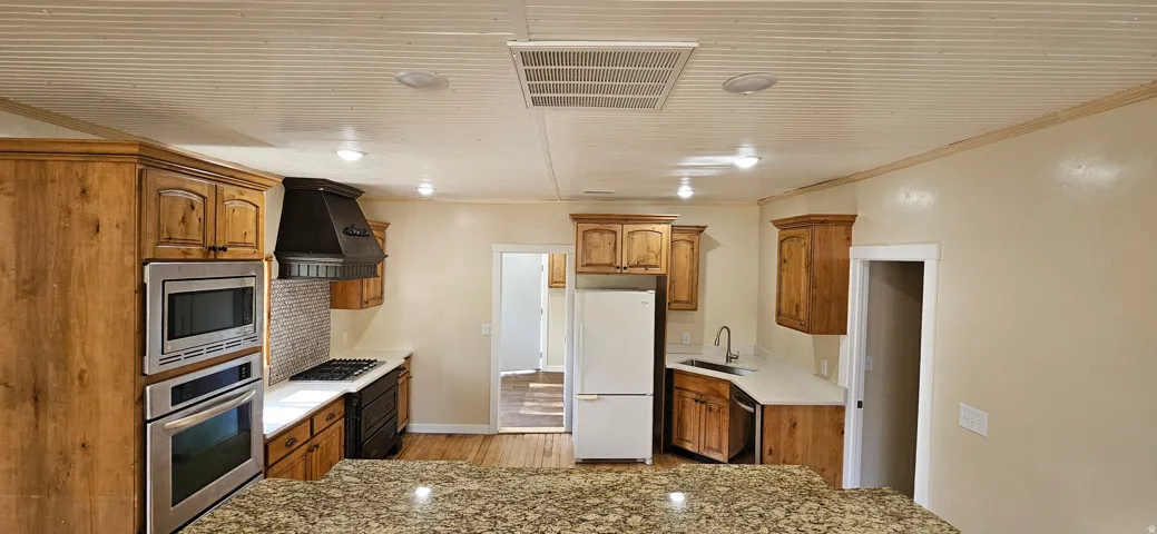 Kitchen featuring a granite countertop island, stainless steel built-in double oven and microwave, dark wood cabinetry, and a textured metal range hood
