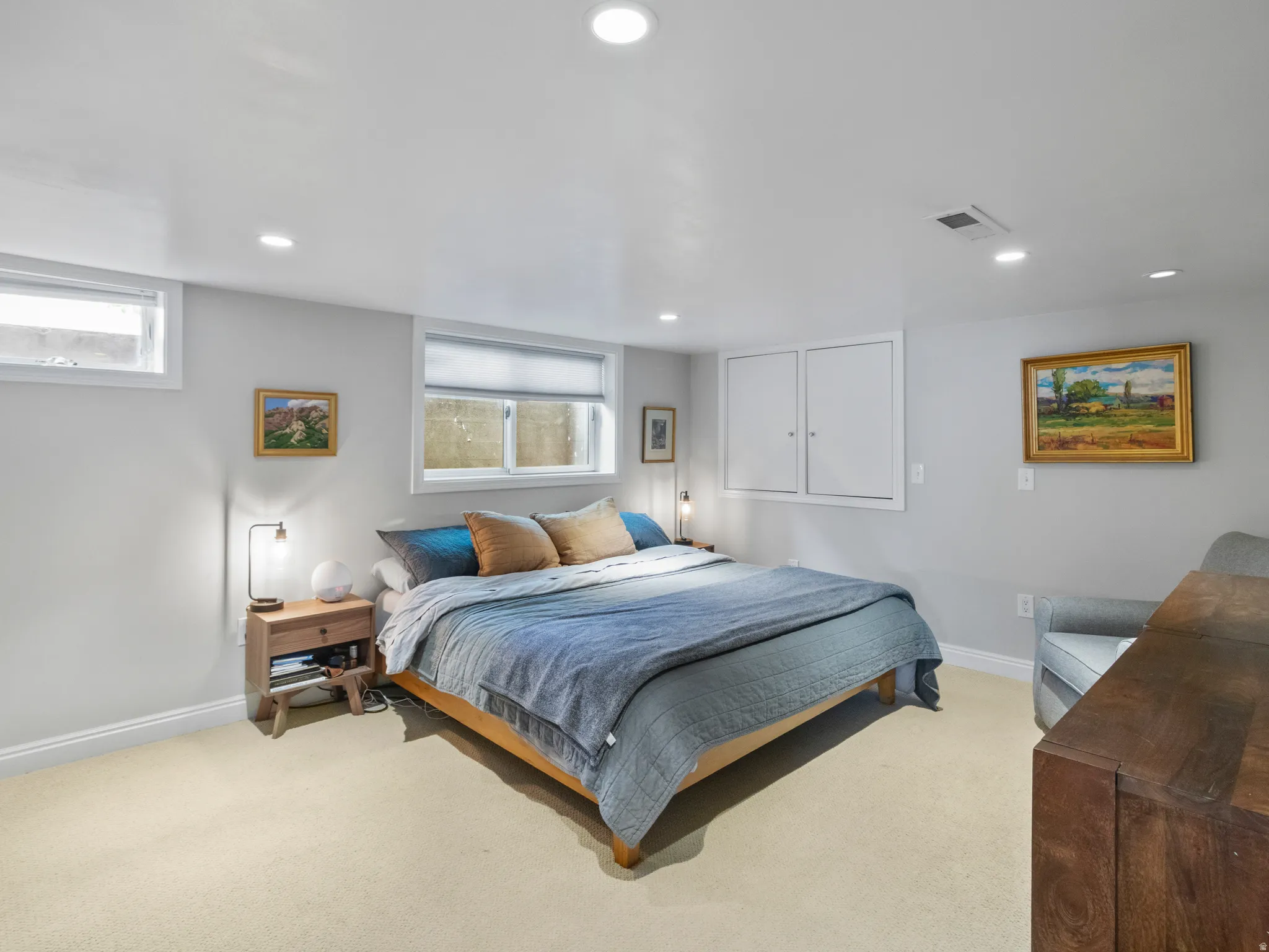 Recessed lighting illuminates this carpeted bedroom featuring two windows with roller shades, white baseboards, and built-in wall cabinetry