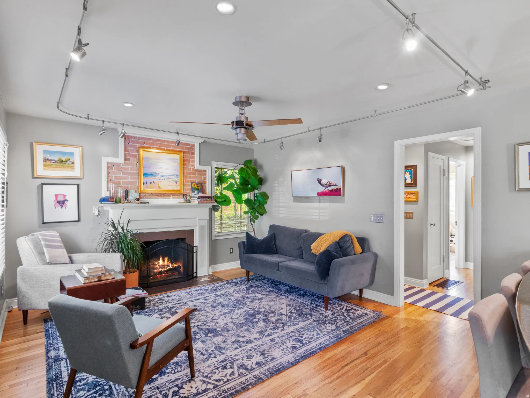 Living room featuring a brick fireplace with a white mantel, hardwood flooring, track lighting, a ceiling fan, and recessed lighting