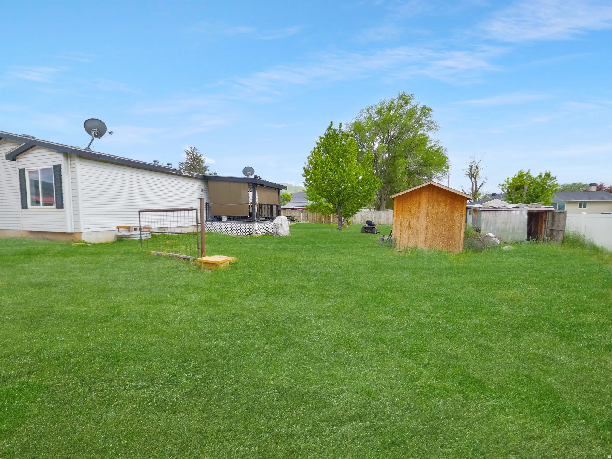 Expansive green lawn with a white vinyl-sided exterior, a covered deck featuring lattice detailing, a freestanding wood shed, and a privacy fence