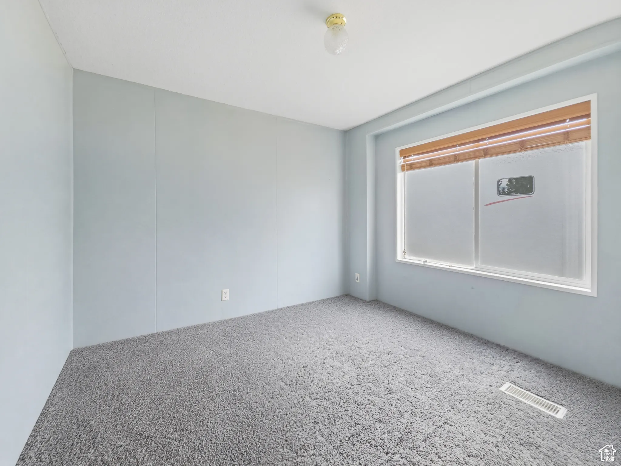 Spacious room featuring light blue paneled walls, gray carpet, a ceiling-mounted light fixture, and a large window with wood-finish blinds