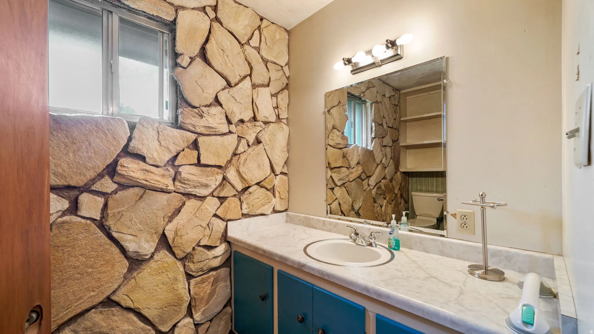 Bathroom featuring an expansive stone wall, a window with horizontal blinds, a vanity with a white countertop and integrated sink, blue cabinetry, and an attached rectangular mirror